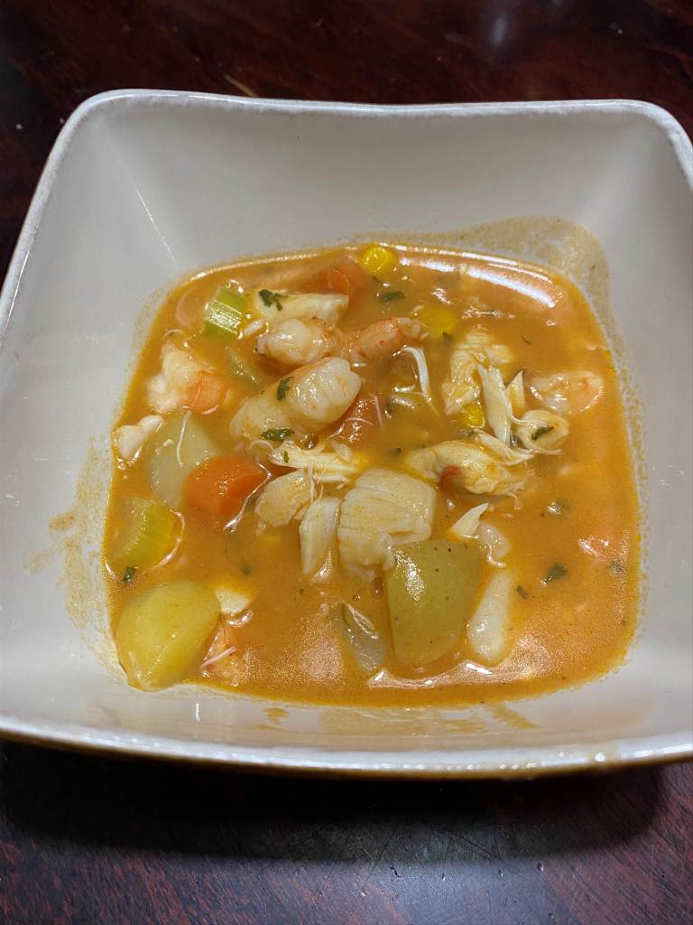 White bowl containing seafood chowder made with shrimp, crab, walleye, and vegetables in a golden broth.