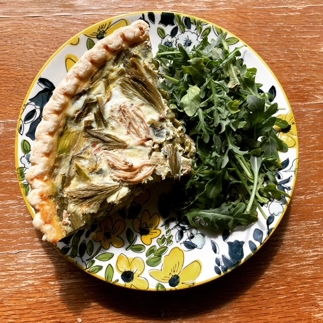 Decorative plate with blue and yellow flowers holding a bundle of fresh greens and a slice of quiche, which is topped with pickled spruce tips and mushrooms.