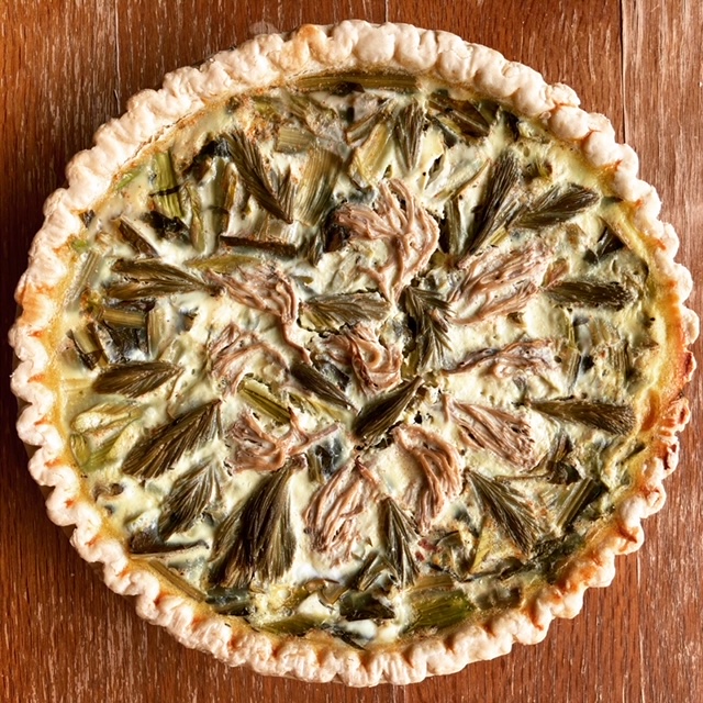 Full quiche set on a wooden backdrop; quiche has an array of pickled spruce tips and mushrooms on top.