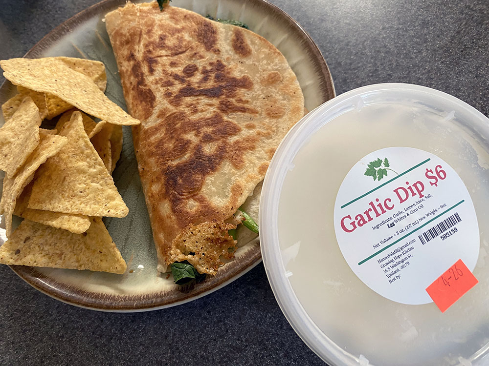 The cooked quesadilla sits on a plate with corn chips next to a tub of locally made garlic dip.