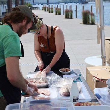 Two people stand over a table at an outdoor cooking demonstration, preparing fish fillets